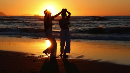 Silhouette of two Latin girls dancing with beers on the beach at sunset during a summer evening