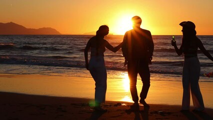 Silhouette of three multiracial friends dancing and drinking beers on the beach at sunset
