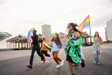 A happy group of people are dancing down the street holding rainbow flags under the colorful sky,...