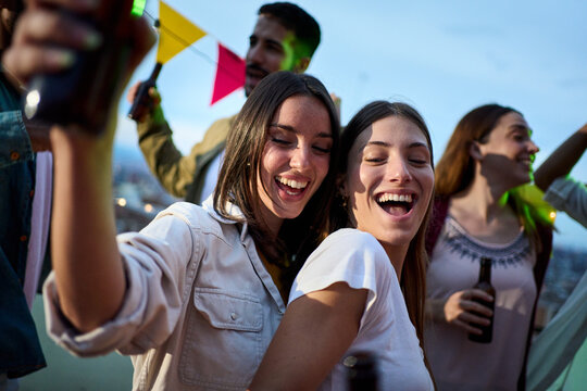 Young cheerful beautiful Caucasian couple women having fun dancing in a party in evening at weekend with friends. Happy female enjoying leisure time in rooftop. Gen z people celebrating summer night - Powered by Adobe