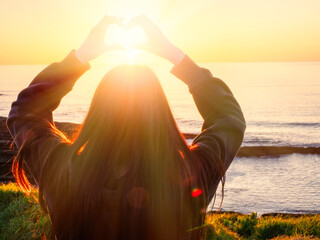 Teenager girl hold hands in heart shape over her head, sunset sun shines through and creates soft...