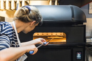 woman with an apron putting the pizza in an electric oven with a peel, working in the restaurant...