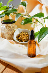 A brown bottle of a natural cosmetic sitting on a natural fiber cloth, on a wooden surface. On the background you can see the ingredients of the product: oats and chamomile, and a green leaved plant.