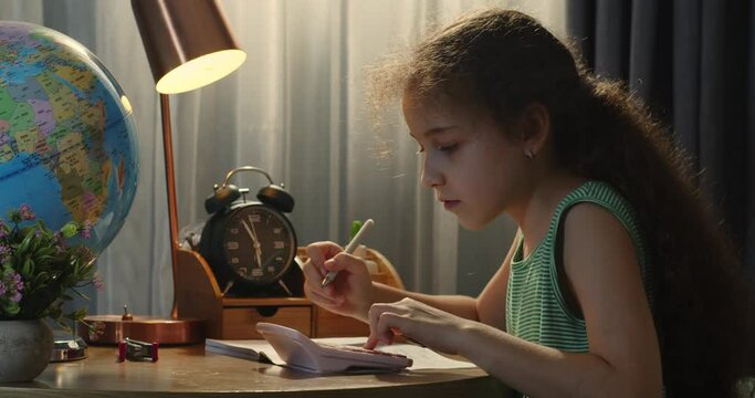 Cute smart primary school child girl learning writing doing math homework sit at home table, adorable pretty little preschool kid studying alone making notes, children elementary education concept