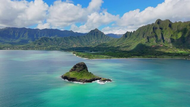 Breathtaking drone shot captures a Chinese hat rock in the midst of blue ocean waters, framed by dramatic green mountain ranges on a sunny day. Hawaii islands, Oahu, Honolulu, USA. 