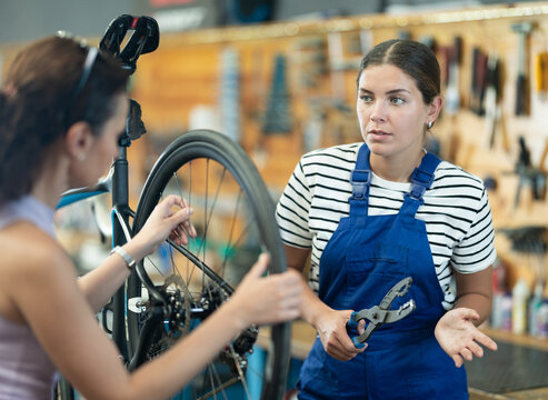 Professional confident young female bicycle technician adjusting bike gear system, fixing sprockets on rear wheel using cassette pliers and explaining repair details to female client in workshop