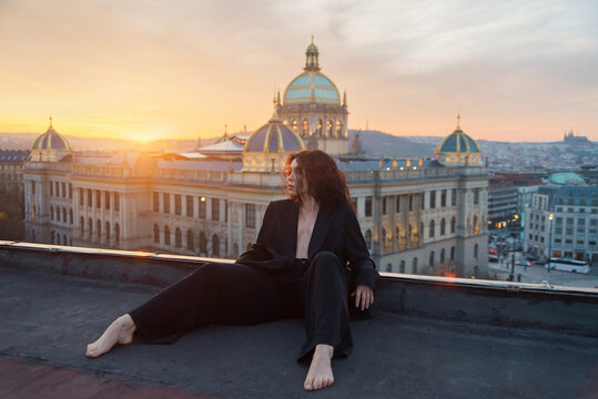 Attractive young curly brunette woman in a black jacket without lingerie sits on a rooftop against a historic building during a cozy sunset