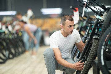Young male customer choosing bike and checking tire on bicycle wheel in modern sport store