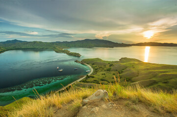 view of the sea surrounded by hills with green grass with the green color of the sea and yellowish shades of the sunset and covered in white clouds on Gili Lawa land © Abdurrahman