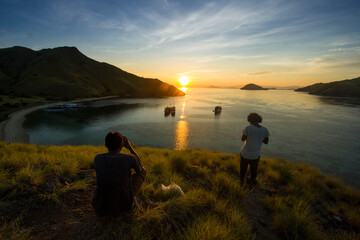 view of the sea surrounded by hills with grass with a dark green sea color and there are 2 people sitting and standing looking at the sunset on Gili Lawa Darat © Abdurrahman