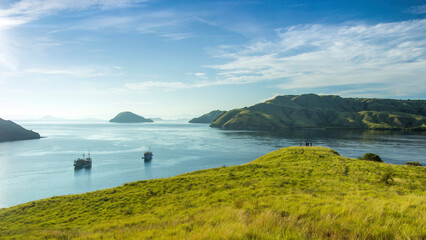 view of the sea and several hills covered with green grass with blue sky and sea and surrounded by white clouds with two sailing ships sailing on Gili Lawa land © Abdurrahman