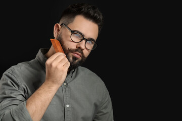 Handsome young man combing beard on black background. Space for text