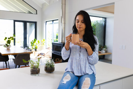 Young biracial woman sitting at home, enjoying coffee in bright kitchen