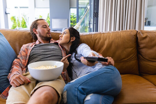 Diverse couple relaxing on couch at home, watching TV and eating popcorn