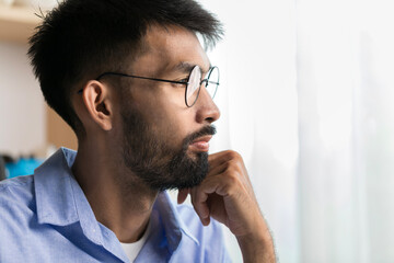 Thoughtful young bearded man sad in eyeglasses looking out window, serious employee thinking about...