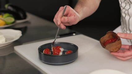 Close up of skillful hands using spoon to drizzling red sauce from small wooden container onto dark plate with pieces of raw meat. Male wearing striped chef attire on kitchen.