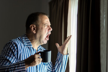 An angry man in striped pajamas with a mug of drink is looking out the window