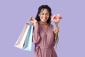 Beautiful young African-American woman with bouquet of gerbera flowers and shopping bags on lilac background. International Women's Day