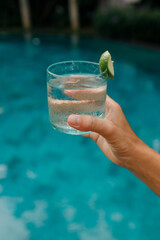 Woman holding refreshing cold drink  water with ice and lime while sunbathing by the pool. Hands close up, vertical shot