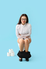 Young Asian businesswoman with bellyache sitting on toilet bowl against blue background