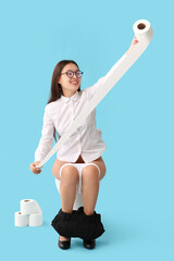 Young Asian businesswoman with paper roll sitting on toilet bowl against blue background