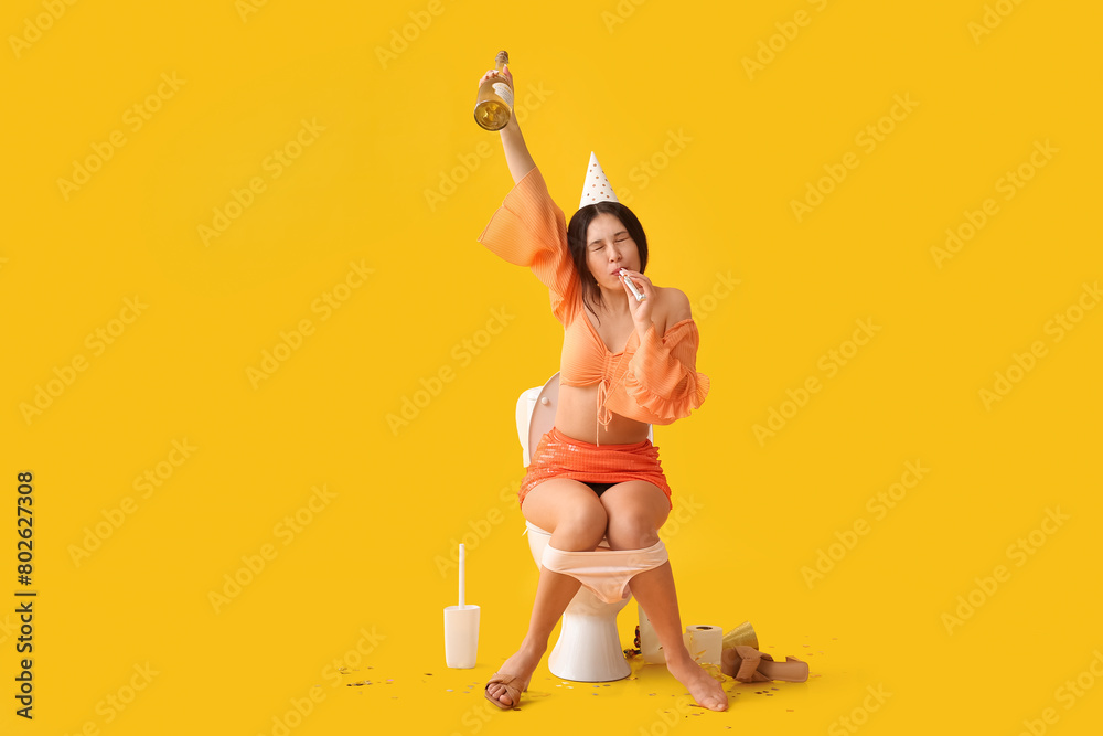 Sticker Young Asian woman with party blower and drink sitting on toilet bowl against yellow background