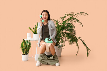 Young Asian woman talking by telephone on toilet bowl against beige background