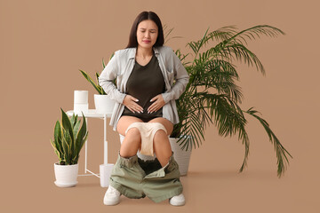 Young Asian woman with cramps sitting on toilet bowl against beige background