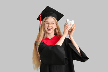 Female graduating student in mortar board with piggy bank on grey background