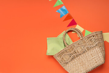 Obraz premium Stylish wicker bag with napkin and flags for Festa Junina celebration on color background