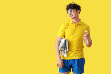 Handsome young happy man with beach blanket and bottle of water on yellow background