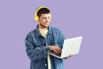 Young bearded man in headphones using laptop on lilac background - obrazy, fototapety, plakaty