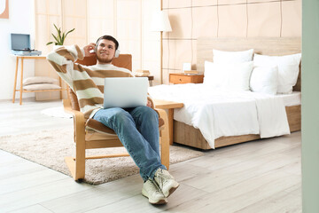 Young bearded man with headphones using laptop on armchair in bedroom