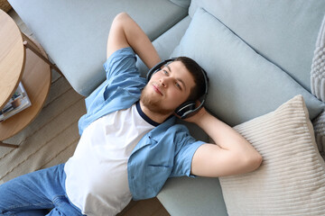 Young bearded man in headphones listening to music beside sofa at home