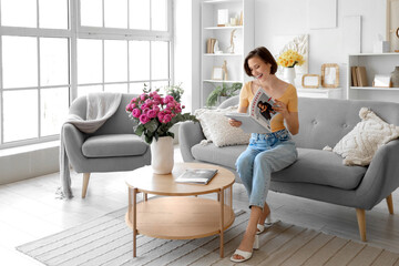 Young woman with pink roses in vase reading magazine at home