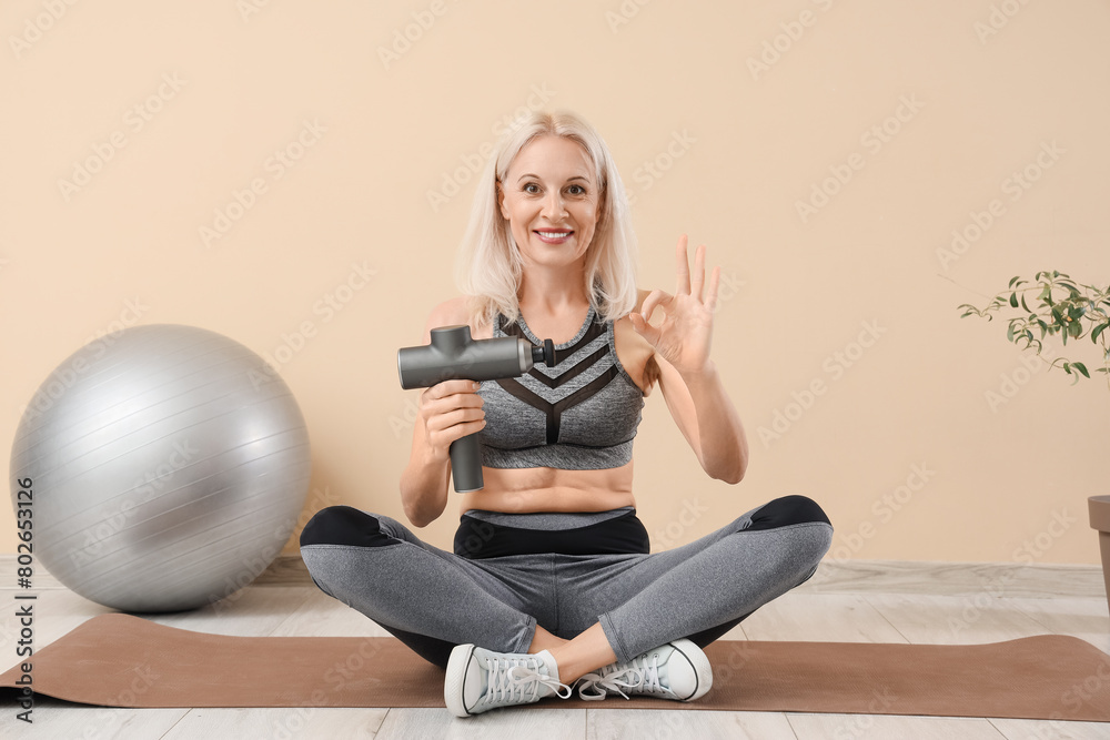 Sticker Sporty mature woman with percussive massager showing OK on mat near beige wall