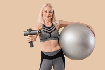 Sporty mature woman with percussive massager and fitball on beige background