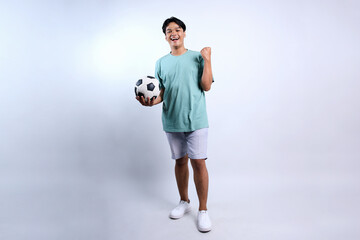 Full Length of Young Asian Man Holding Soccer Ball Clenching Fist Celebrating Victory Isolated On...