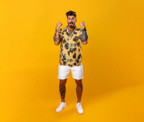 Fototapeta premium Latino man celebrating, in a yellow shirt with patterns and white shorts, tattoo in his arms, standing on a yellow background. 