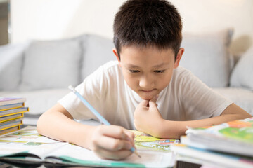 Asian little kid boy concentrating on studying,writing and reading a book,serious focused student doing homework on desk at home,elementary child schoolboy eager to learn,education,back to school.