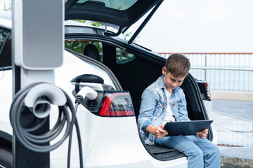 Little boy sitting on car trunk, using tablet while recharging eco-friendly car from EV charging...