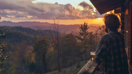 A young man in a flannel shirt leans on the railing of the porch of a rustic cabin overlooking forests and mountains at sunset near Oak Ridge, Tennessee. 
