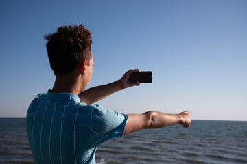 A young attractive guy is posing while relaxing on the sea and taking a photo with his phone.