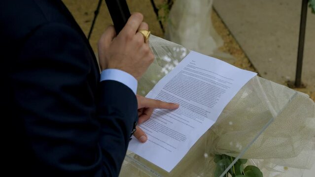 a man reads a text on a transparent plexiglass lectern at a wedding, it's a speech, focus on the sheet and the finger running over the lines, the hand holding the microphone