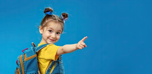 A little girl with a large school backpack on a blue background points her finger at an empty space. Copy space.