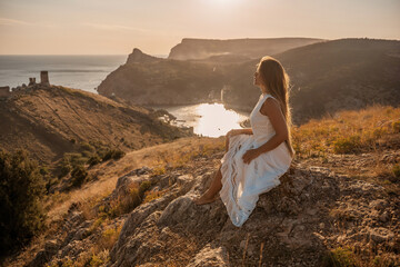 A woman in a white dress sits on a rock overlooking a body of water. The scene is serene and...