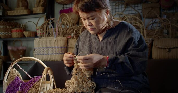 Slow motion scene while an Asian female artisan who is a local villager knitting folk basket made from leftover paper band to sell as extra income in the basketry shop shop.