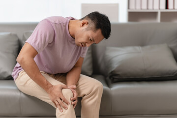 Knee pain sick body health care concept A young man has knee pain, sitting on the sofa at home, touching his leg, feeling pain in his knee.