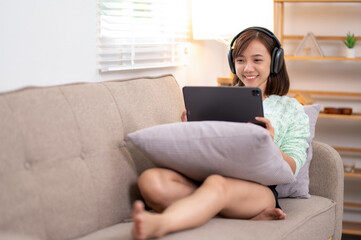 A woman is sitting on a couch with a tablet. Wearing headphones and enjoying her music.