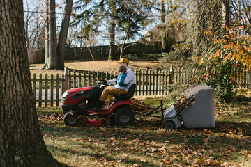 Father drives lawnmower with toddler collecting leaves in fall